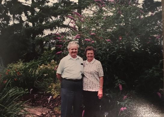 Elderly couple stands together in a lush garden, enjoying a sunny day filled with blooming flowers.