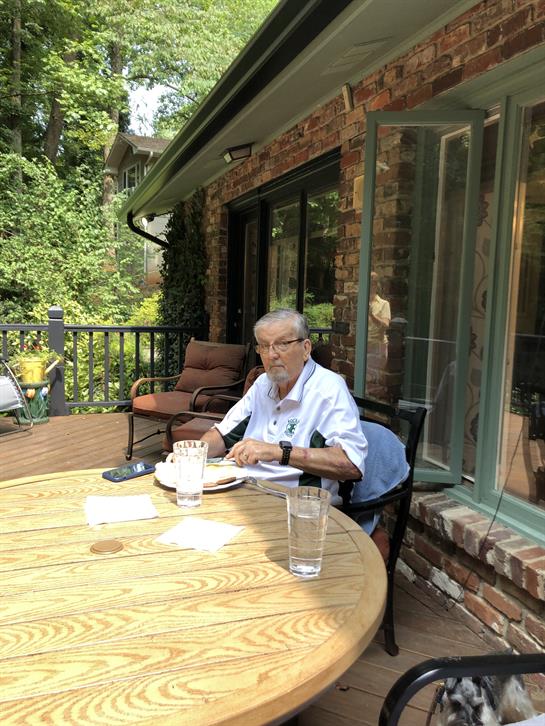 Senior individual seated at a wooden table enjoying a meal while surrounded by lush plants.
