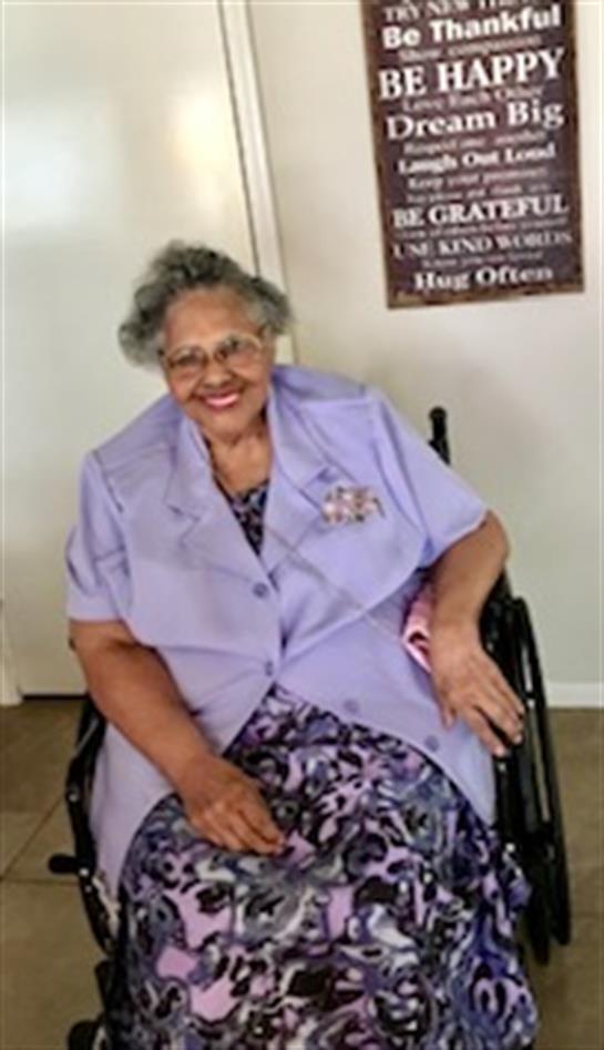 A joyful elderly woman in a wheelchair with glasses smiles while wearing a pastel outfit.