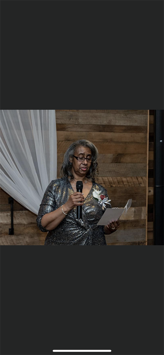 A woman speaks to guests at a decorated venue, holding a microphone and reading from a card.