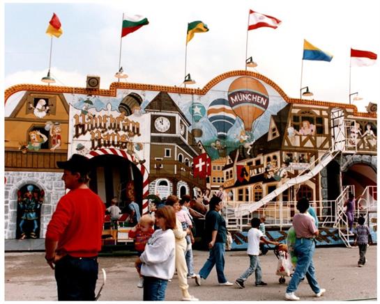 Families wander a vibrant fairground with colorful attractions and international flags.