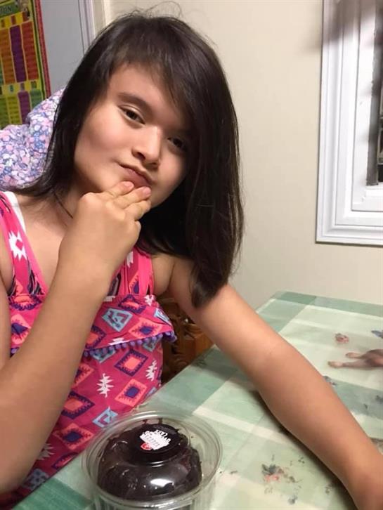Girl with dark hair wearing a pink dress sits at a table, thoughtfully enjoying dessert and posing.