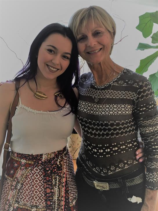 Two women stand close together, smiling warmly, surrounded by greenery and light.