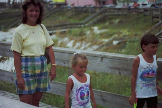 Two adults and a child stand together on a boardwalk by the beach on a sunny afternoon.