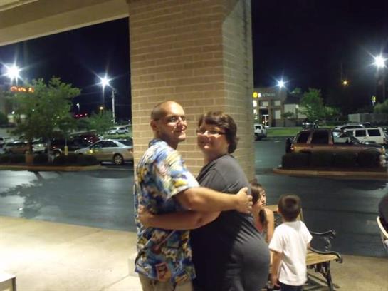 A couple stands happily embracing each other outside a busy shopping center at night.