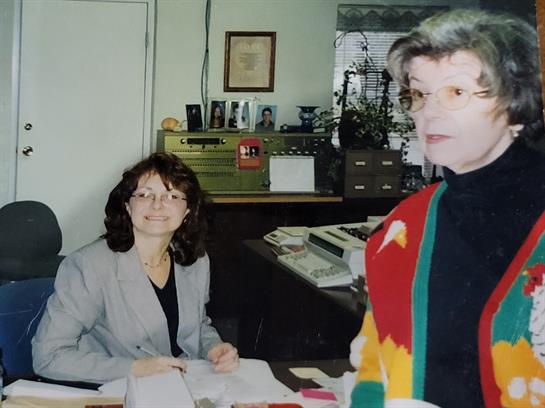 Two women talk in an office filled with paperwork and equipment, building a friendly rapport.