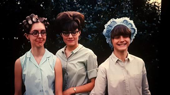 Three women stand together smiling, showcasing their unique, retro hairstyles and glasses.