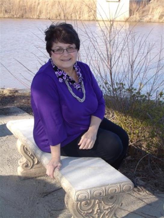 A woman with glasses enjoys a sunny day sitting on a stone bench near a lake, surrounded by nature.