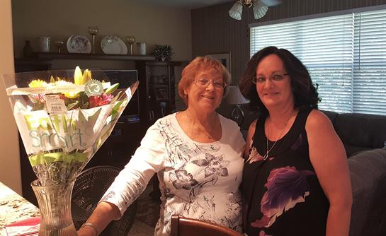 Two women smile joyfully while posing by a table adorned with fresh flowers indoors.