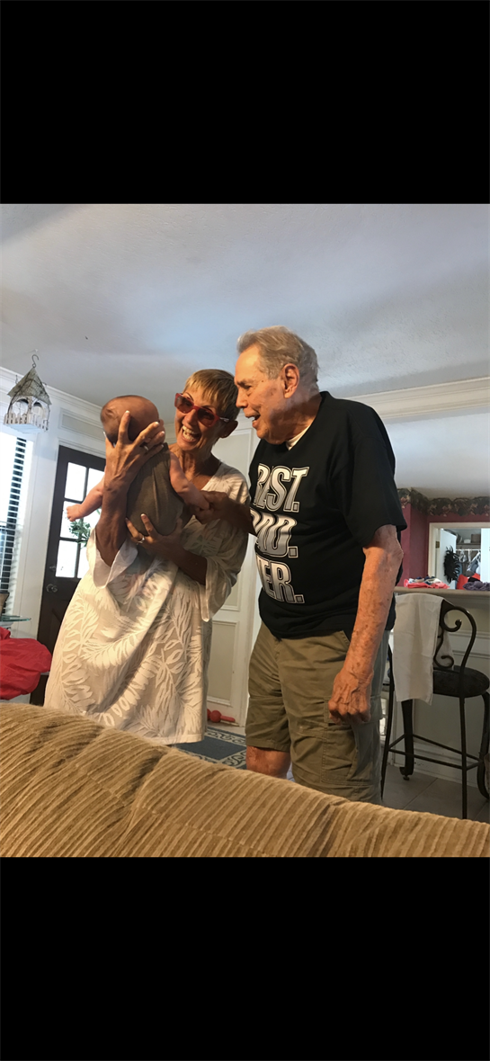An elderly couple smiles and plays with a baby, sharing a warm moment in a comfortable home.
