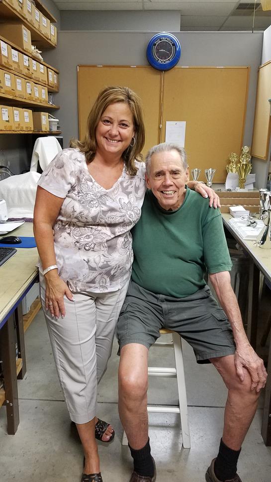 A woman stands alongside an elderly man sitting on a stool in a cozy workshop, both smiling.