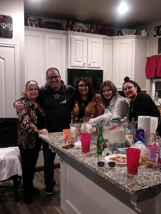 A group of five friends stands together in a warm kitchen, enjoying drinks and smiles.