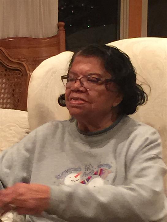 An older woman wearing glasses smiles as she shares stories in a cozy living room setting.