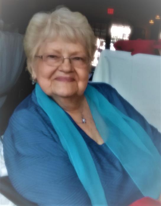 An elderly woman with glasses smiles warmly, wearing a turquoise scarf.