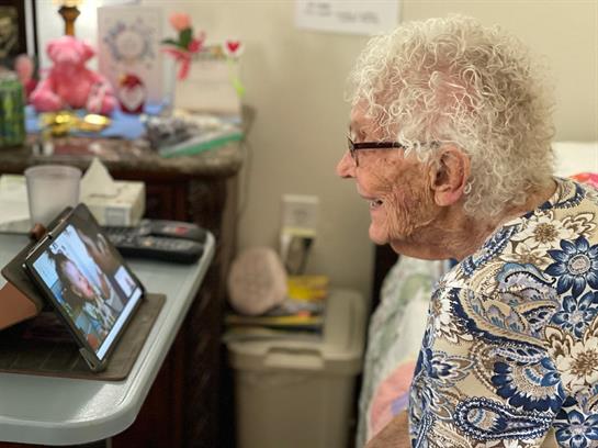 An elderly woman is smiling widely while engaged in a video call, enjoying a moment with loved ones.