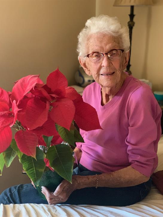 An elderly woman sits with a vibrant red flower arrangement, smiling warmly indoors.