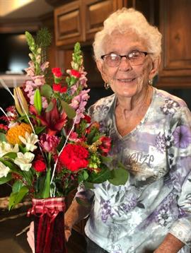 Elderly woman smiles while holding a colorful bouquet of flowers in a cozy kitchen setting.
