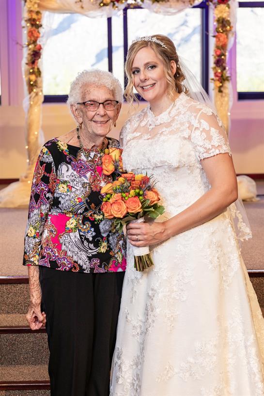 A bride proudly stands with her smiling grandmother amid wedding decorations.