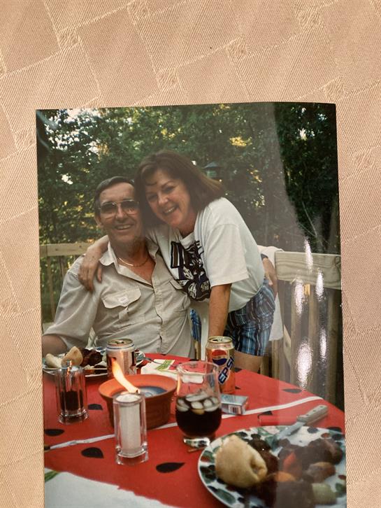 A man and a woman share a happy embrace at a cheerful outdoor gathering, surrounded by drinks.