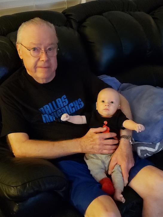 A grandfather sits on a couch, holding a lifelike baby doll, both looking relaxed and content.
