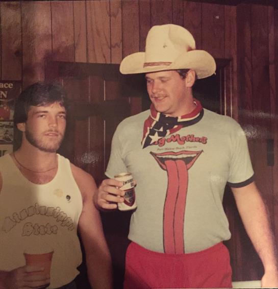 Two men socialize while holding drinks in a rustic bar decorated with wooden panels.