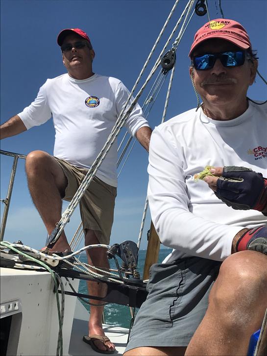 Two sailors relax on a sailboat under a bright sky, enjoying fishing and the beautiful weather.