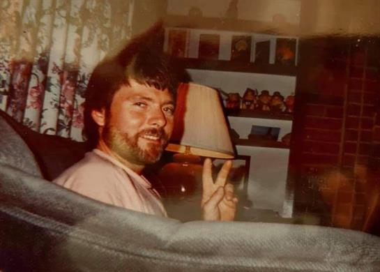 A man smiles and shows a peace sign while relaxing in a chair, surrounded by warm indoor decor.
