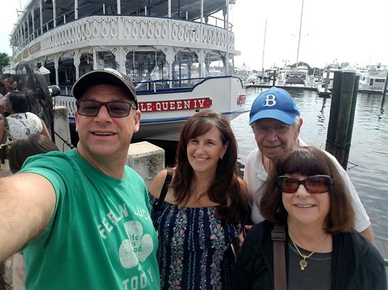 Friends pose happily at the harbor by a riverboat on a sunny day with boats in the background.