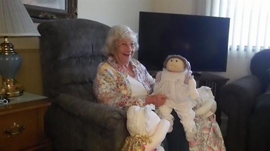 An elderly woman relaxes in a chair with a doll and her handmade creations around her.