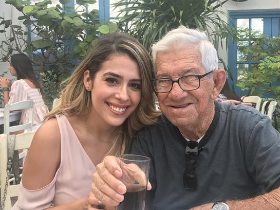An elderly man smiles with a young woman, both enjoying drinks in a lively setting.