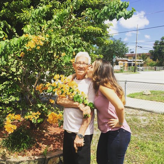 A woman kisses her grandfather on the cheek while they stand among vibrant yellow flowers.