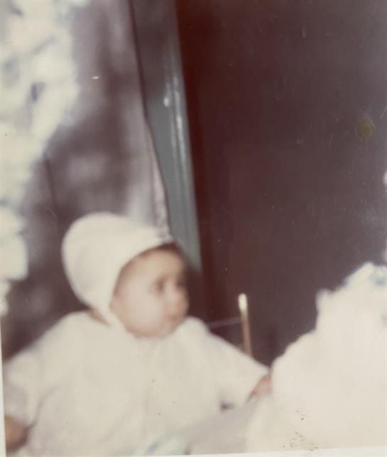 A baby in a white outfit and hat sits with a family member, capturing a warm memory indoors.