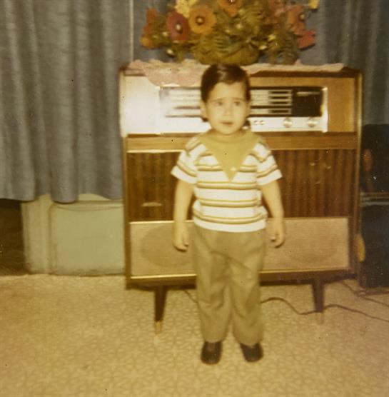 A young child poses in a living room with a retro radio and blue curtains nearby.