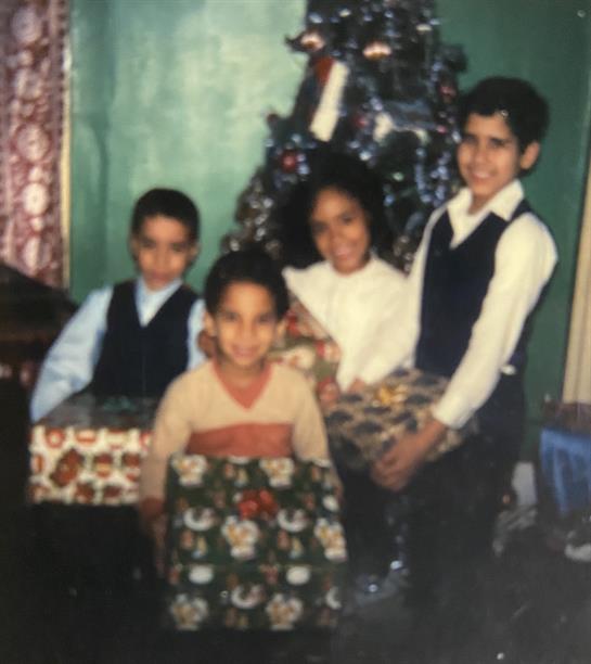 Four kids joyfully gather by a Christmas tree, holding colorful wrapped gifts.