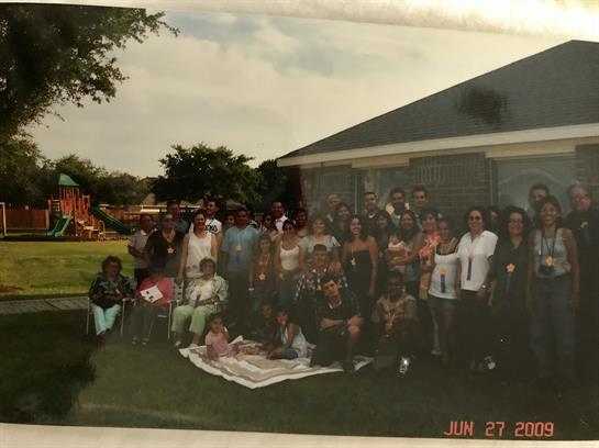 Large group of people smiling and sitting on a grassy area during a sunny day in a park.