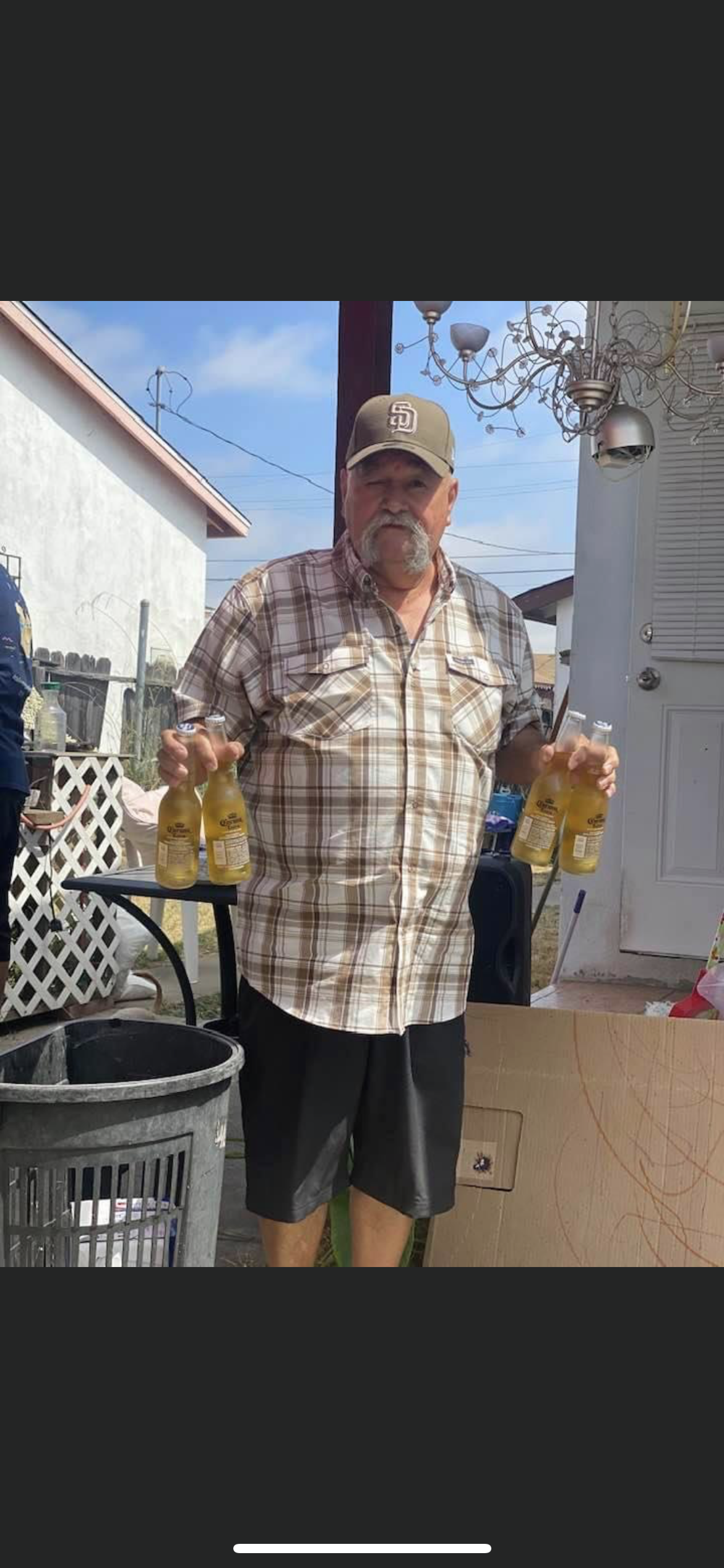 A man stands in a backyard, carrying multiple bottles while a barbecue is set up nearby.