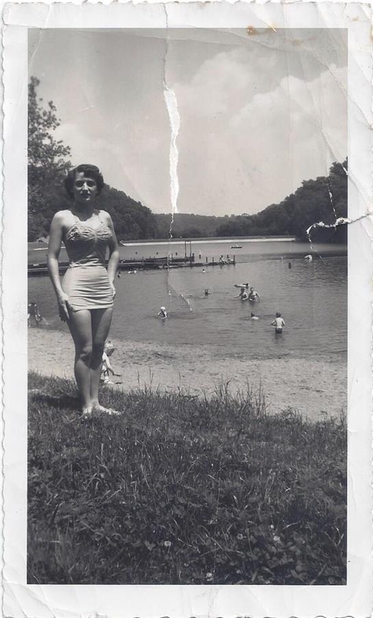 A young woman in a swimsuit poses at the edge of a lake on a sunny summer day.