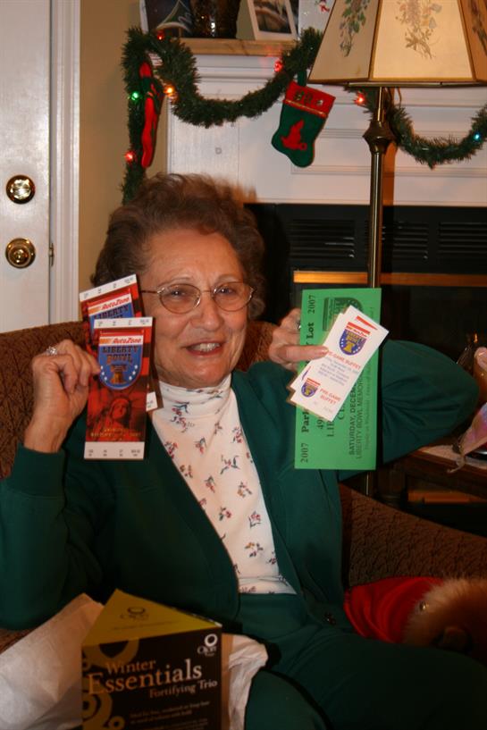 A cheerful elderly woman holds up tickets and envelopes, surrounded by holiday decorations indoors.