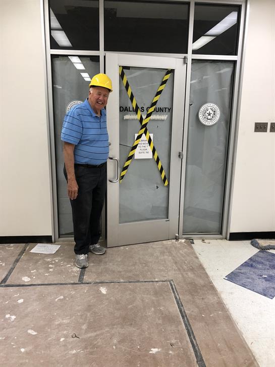 A man in a yellow hard hat stands near a construction door marked with caution tape.