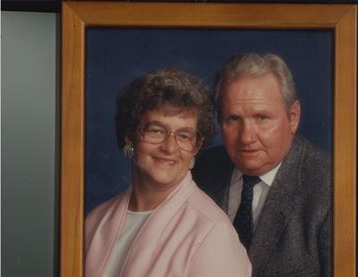 An older couple stands closely together, smiling, in a formal portrait setting with soft lighting.