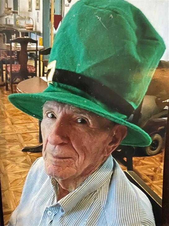 An elderly man wearing a vibrant green hat smiles while sitting in a lively venue with friends.
