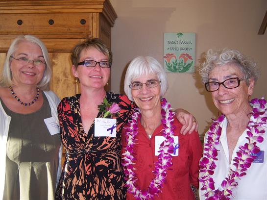 Four women smile and pose together at a joyful gathering, wearing floral leis and name tags.