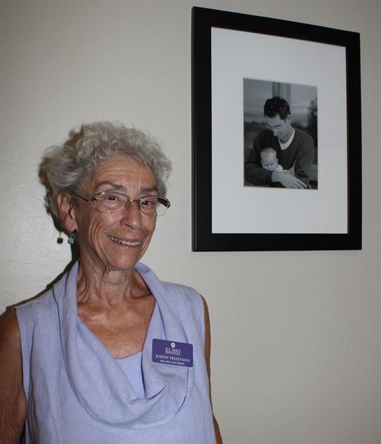 Elderly woman with glasses smiles while standing next to a framed photo on the wall.