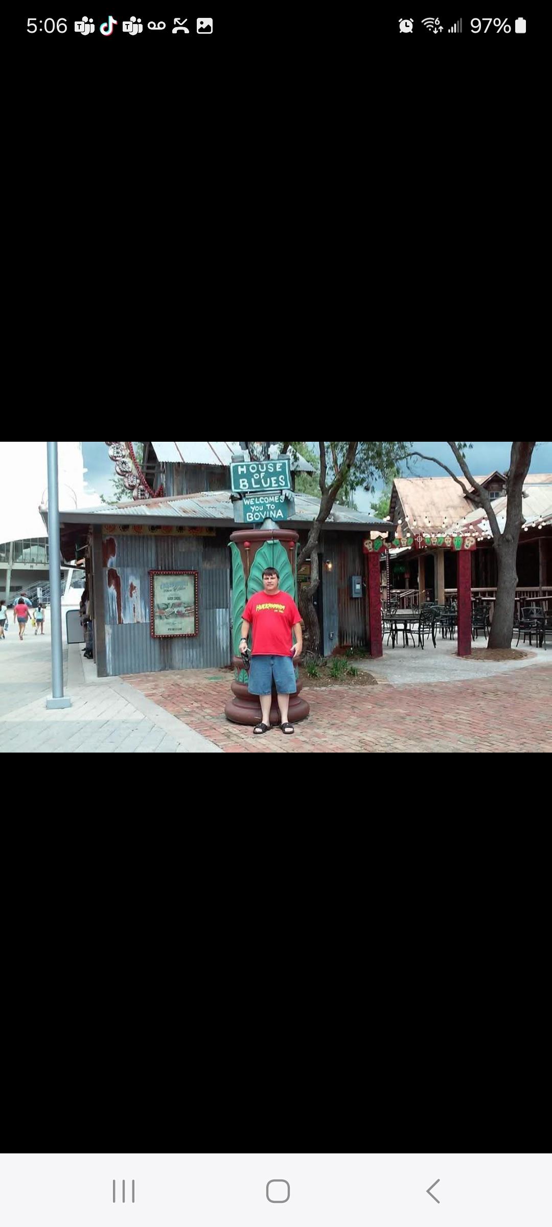A person in a red shirt stands by a colorful storefront on a sunny day in a bustling location.