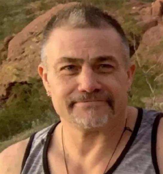A man poses with a slight smile in a rocky outdoor setting under clear blue skies.