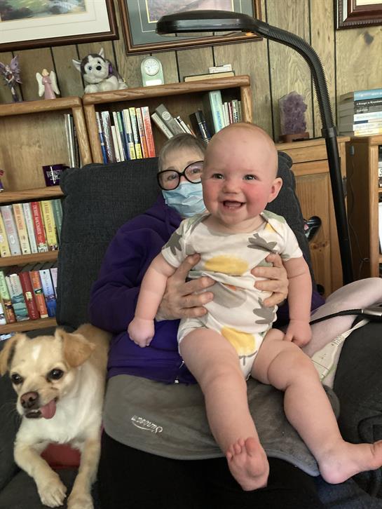 A joyful baby sits on a person's lap with a dog nearby, all relaxing in a cozy living room.