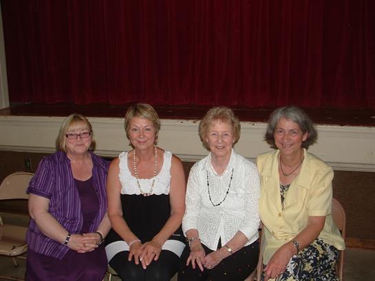 Four women sit side by side, dressed elegantly, smiling at a formal gathering in a theater.
