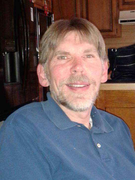 A man is joyfully smiling while seated indoors, surrounded by warm wooden decor and soft lighting.