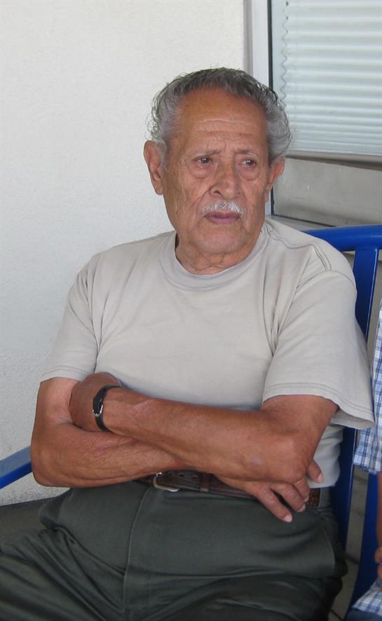 An elderly man sits relaxed with crossed arms, wearing a light shirt in a casual environment.