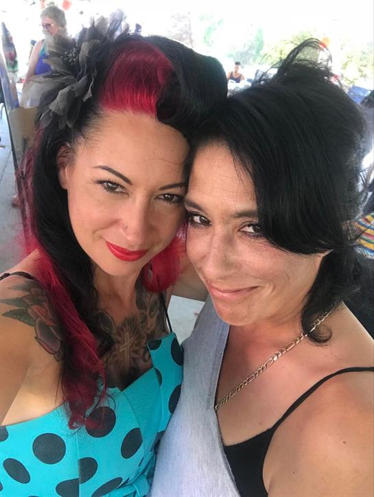 Two women smile and pose for a joyful selfie at a lively outdoor event on a sunny day.